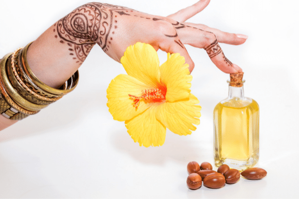 Ženska ruka s nizom narukvica i tetovažom drži žuti cvet i dodriuje bočicu sa uljem pored plodova jojobe